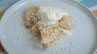 A section of the Banana Coconut Cobbler, served on a white plate topped with shaved coconut and ice cream, which has melted slightly and runs down into the cobbler's nooks and crannies.